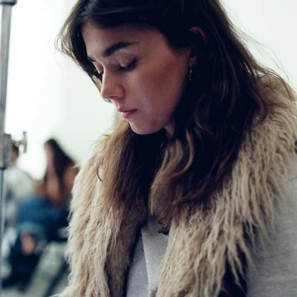 A woman with long brown hair, wearing a beige faux fur vest and grey sweatshirt, looks down indoors in a medium portrait shot.