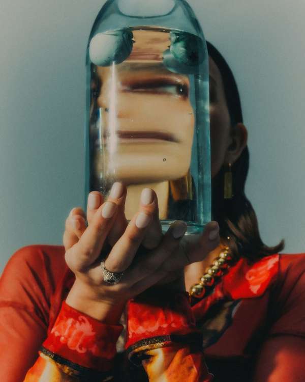 Woman in red top holds a glass bottle filled with water, distorting her face, with a soft light and a neutral wall in the background.