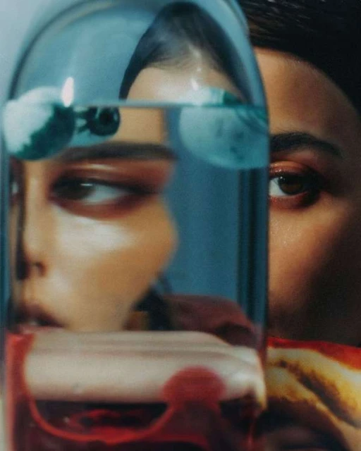 Two adult females are partially visible behind a glass bottle of red liquid in an indoor setting, artistic lighting.
