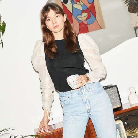 A young woman with brown hair wearing a black top with sheer puff sleeves and light wash jeans holding a patterned mug.