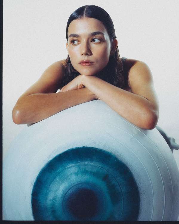 Woman with dark hair leans on large eyeball in a photo studio. Moody lighting highlights her face and the ball's blue iris.