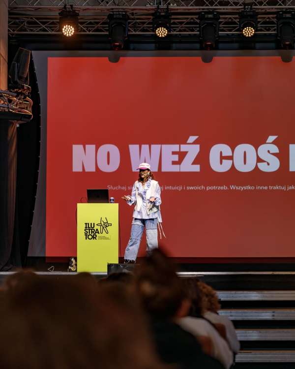 Woman presents on stage to audience at event. The presenter stands near a yellow stand with a laptop. Red backdrop has white text.