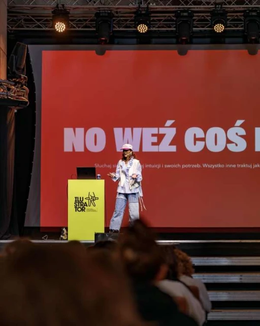 Woman presents on stage to audience at event. The presenter stands near a yellow stand with a laptop. Red backdrop has white text.
