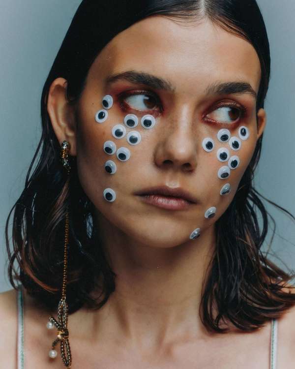 Close-up shot of a young adult woman with many googly eyes glued to her face, wearing a spaghetti strap tank top and statement earrings.