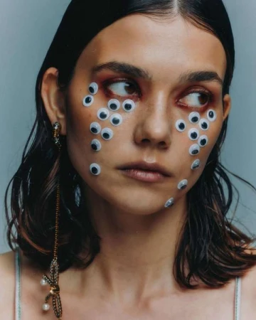 Close-up shot of a young adult woman with many googly eyes glued to her face, wearing a spaghetti strap tank top and statement earrings.