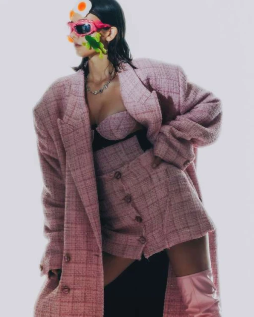 Woman in a pink tweed skirt suit and matching boots poses against a neutral background with fashion-forward, artistic accessories.