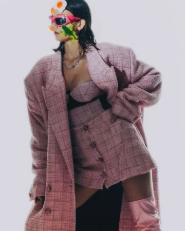 Woman in a pink tweed skirt suit and matching boots poses against a neutral background with fashion-forward, artistic accessories.