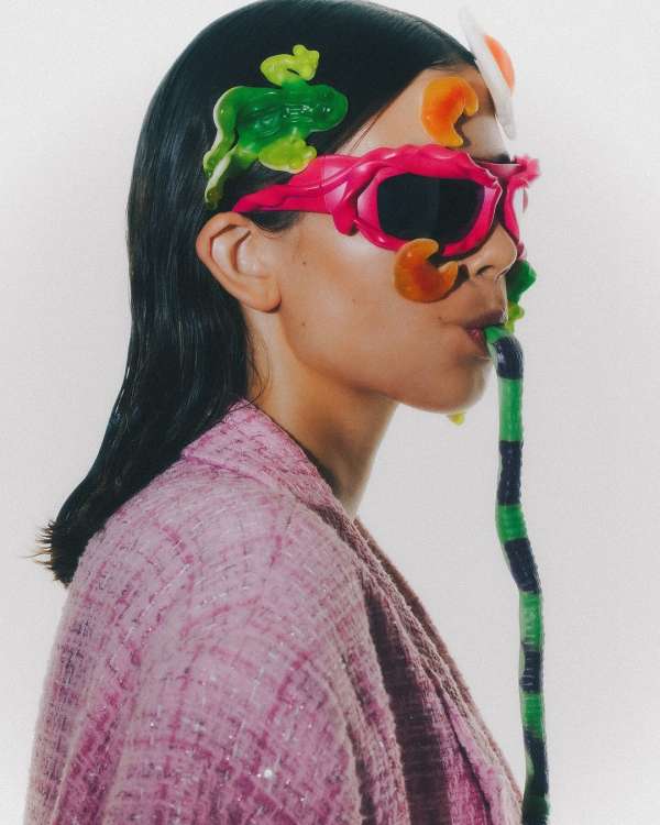 Woman in pink tweed jacket wearing pink sunglasses and playful candy accessories in a studio shot, against a solid white backdrop.