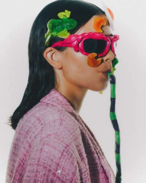 Woman in pink tweed jacket wearing pink sunglasses and playful candy accessories in a studio shot, against a solid white backdrop.