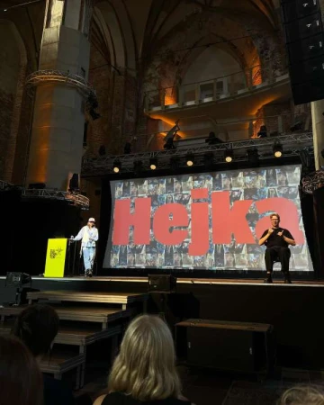 Two presenters on a stage in front of a large screen, addressing an audience; stage lighting and architectural details visible.