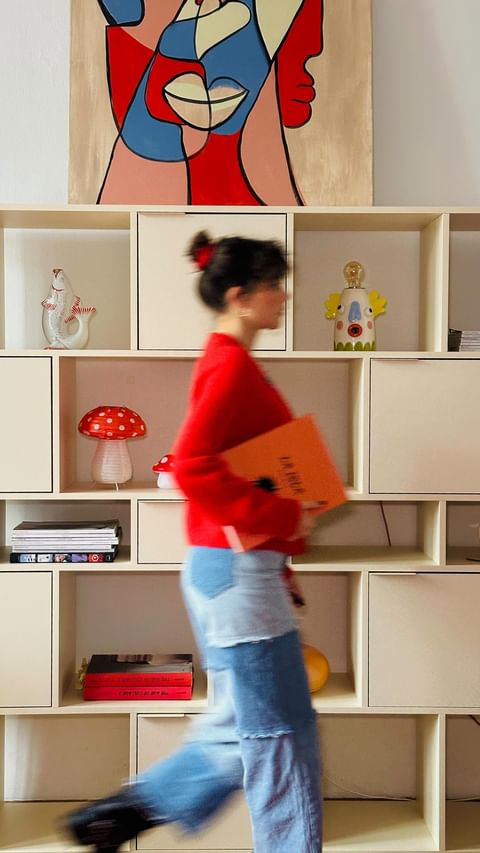 Woman in red sweater walks past a cream-colored shelving unit with art, books, and decorative objects in a brightly lit interior space.