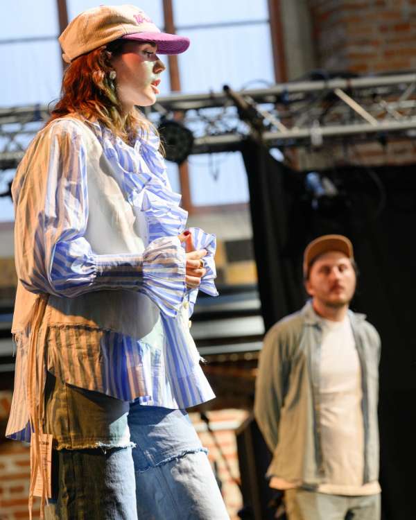 A woman stands on a stage wearing a cap, blue and white ruffled shirt, and jeans, while a man stands in the background wearing a cap.