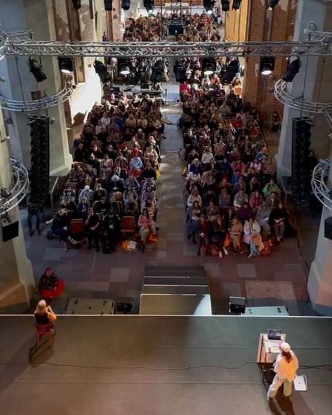 Large audience seated indoors at a conference, watching presenters on stage with lighting rigs overhead in a large hall with brick walls.