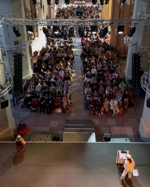 Large audience seated indoors at a conference, watching presenters on stage with lighting rigs overhead in a large hall with brick walls.