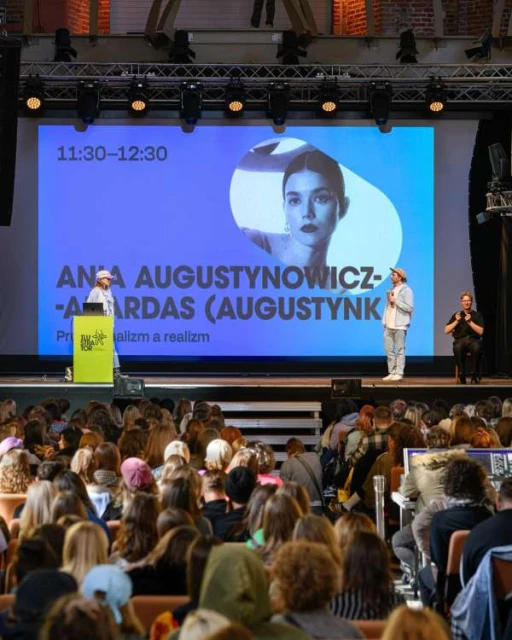 Large group of people at an indoor event, with two speakers on stage in front of a screen displaying a portrait image and presentation text.