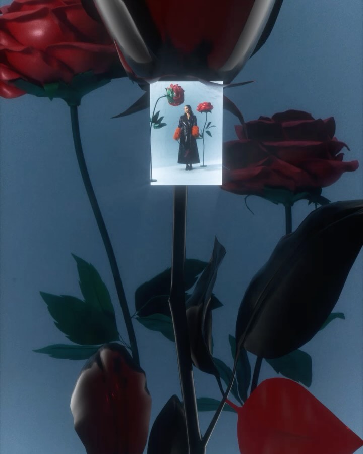 A woman in a dark patterned coat poses in a photo booth-like frame with large red rose props against a pale blue background in studio lighting.