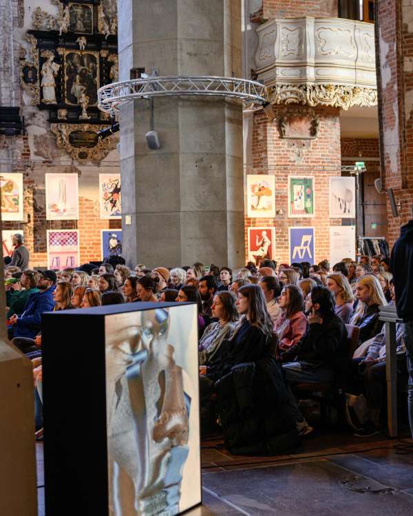 Large group of people sit in rows at presentation with large screen showing an abstract, textured image in a high-ceilinged, ornate hall.