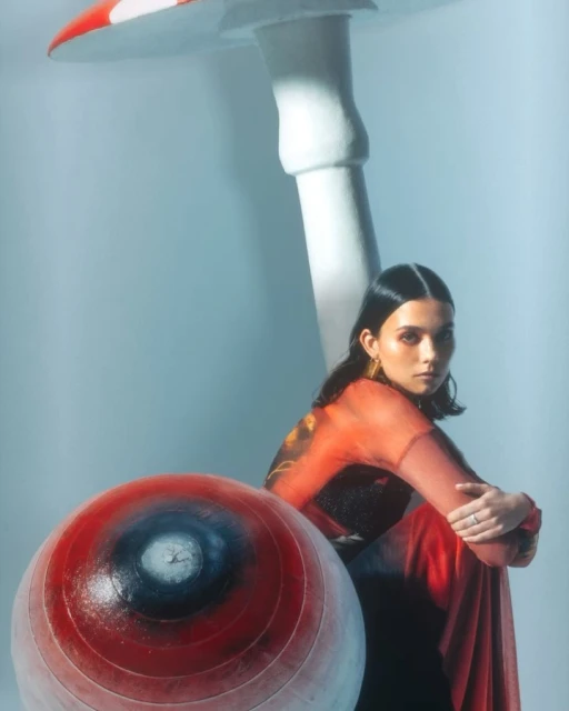Brunette woman poses seated next to a large mushroom and a red-and-white spherical object in a brightly lit photo studio setting.