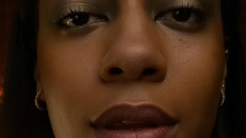 Close-up portrait of an adult woman with warm skin tone, featuring her face and minimal jewelry against a dark, blurred background.