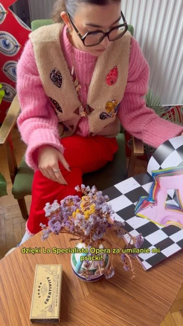 Woman in a pink sweater and vest sits at a table with flowers and a book featuring colorful graphics and checkerboard pattern.