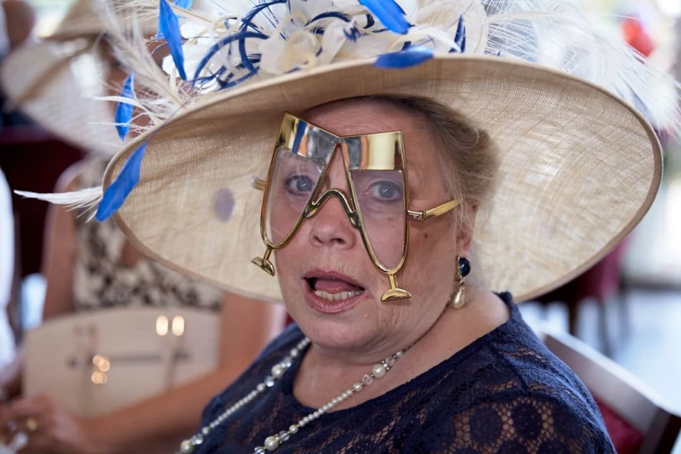 Woman with champagne-glass novelty glasses and a wide-brimmed feathered hat smiling at an event, wearing pearl jewellery.
