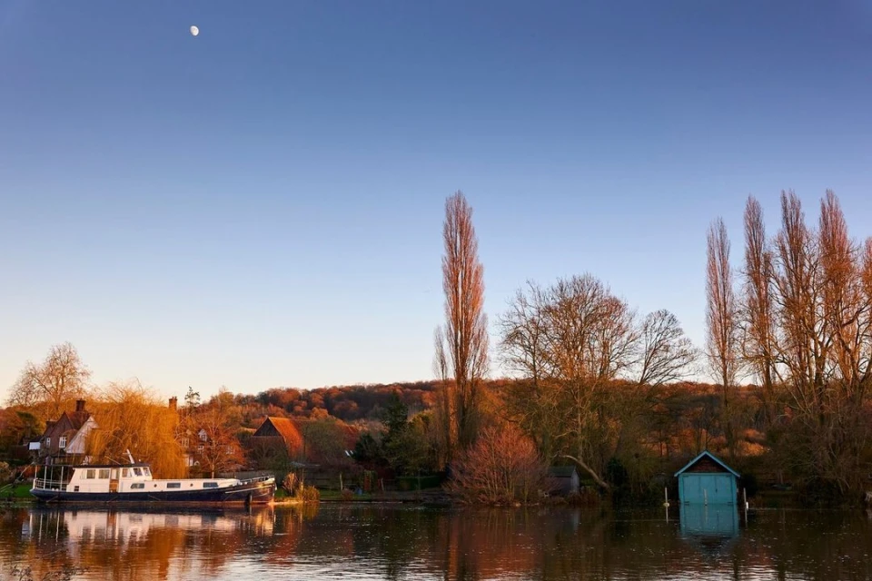 Scenic view of a river with a boat, trees, and houses along the bank under a blue sky with a visible moon at daytime.