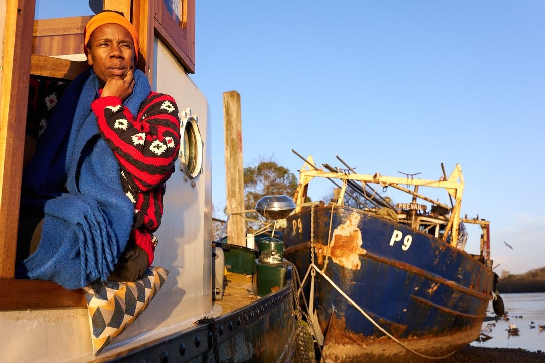 Adult sits on a boat seat near another vessel wearing a bright orange hat, patterned sweater, and blue scarf, looking thoughtful outdoors.