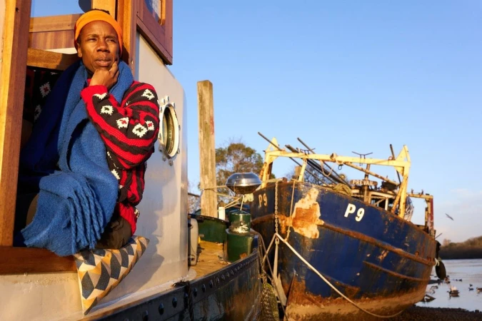 Adult sits on a boat seat near another vessel wearing a bright orange hat, patterned sweater, and blue scarf, looking thoughtful outdoors.
