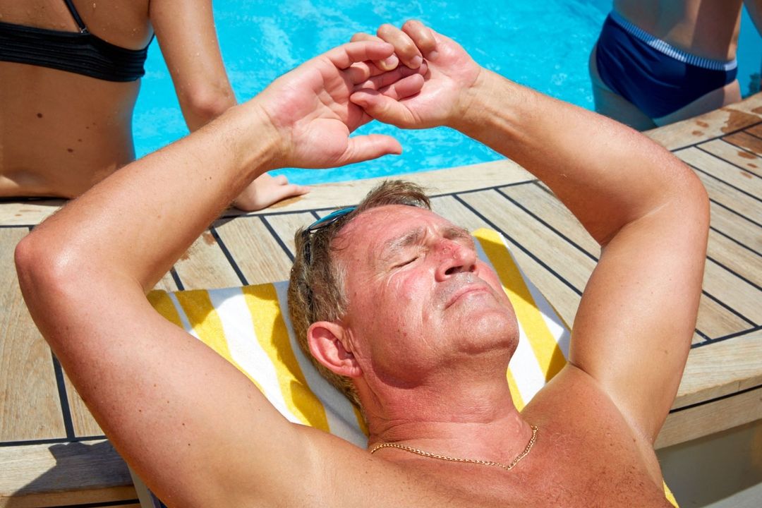 Man sunbathing on a yellow and white striped towel by a pool, with two other people partially visible in the background.