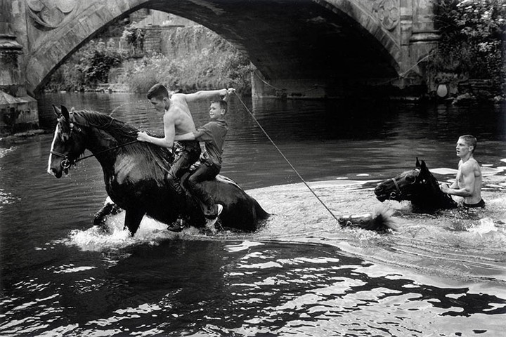 Black and white shot of two men riding a horse through water, with a third man holding a rope attached to a second horse.