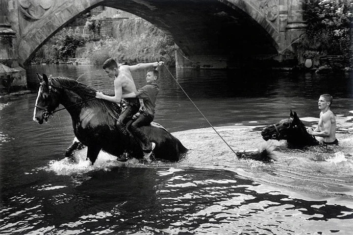 Black and white shot of two men riding a horse through water, with a third man holding a rope attached to a second horse.