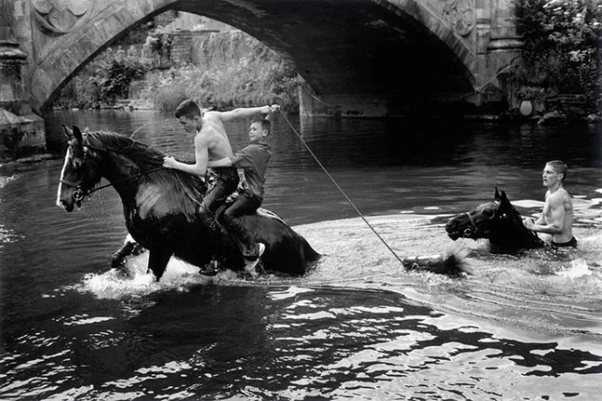 Black and white shot of two men riding a horse through water, with a third man holding a rope attached to a second horse.