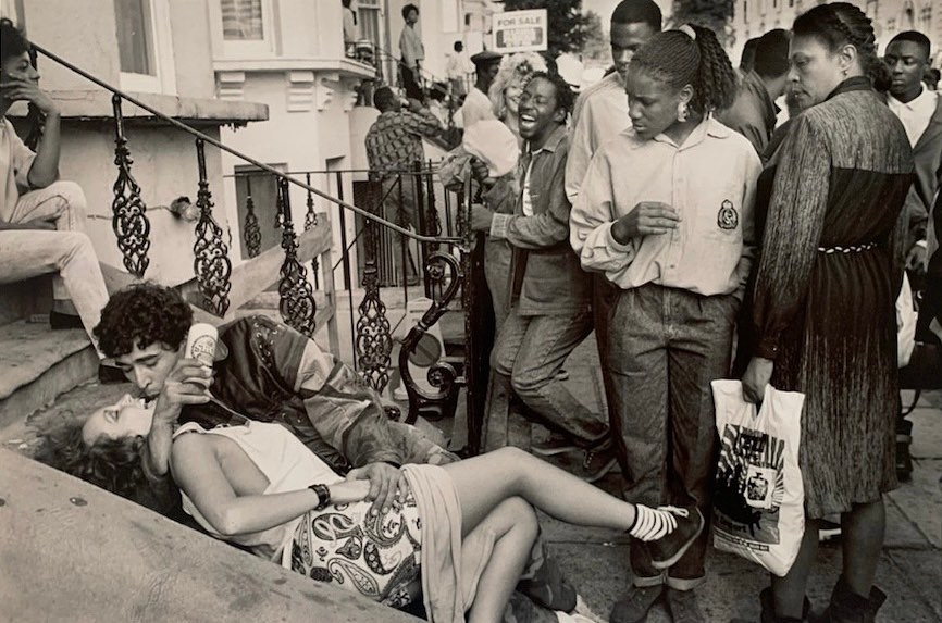 Monochrome photograph captures an embracing couple on steps, surrounded by a crowd, creating a candid, street photography moment.