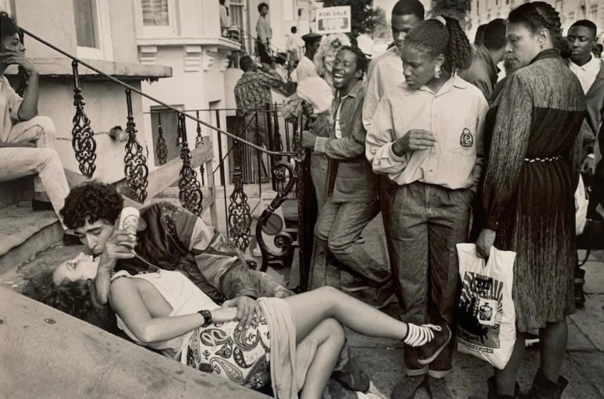 Monochrome photograph captures an embracing couple on steps, surrounded by a crowd, creating a candid, street photography moment.