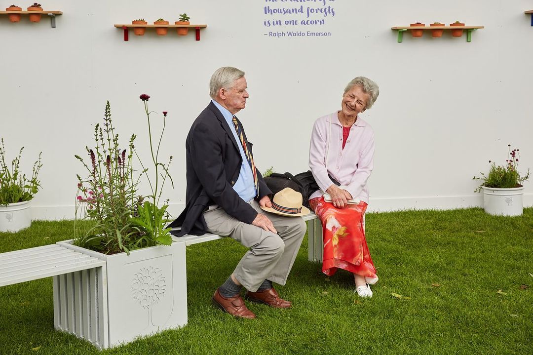 Elderly couple sitting on a bench amidst greenery, smiling at each other on a sunny day, with a white wall in the background.
