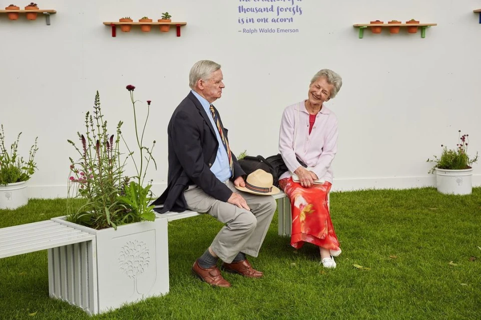 Elderly couple sitting on a bench amidst greenery, smiling at each other on a sunny day, with a white wall in the background.