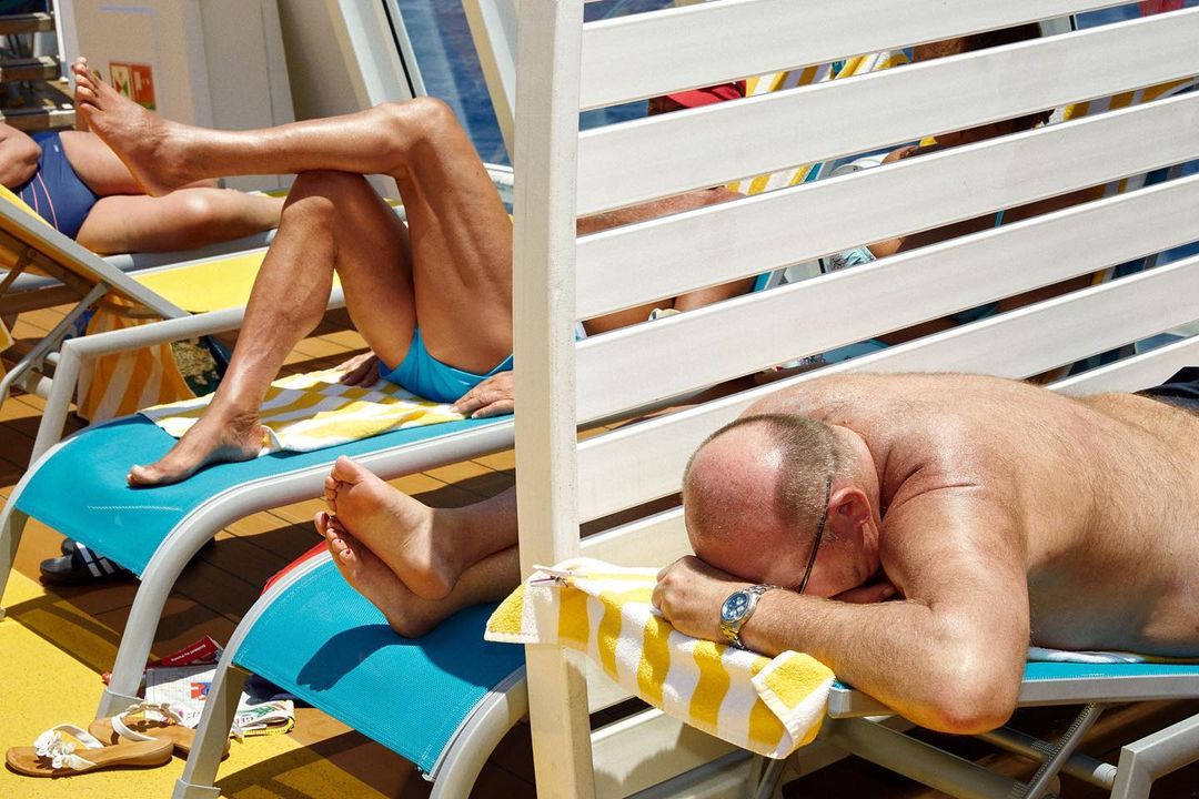 Men sunbathing on deck chairs in bright sunlight near the ocean, wearing swimsuits, with a white partition in an outdoor environment.
