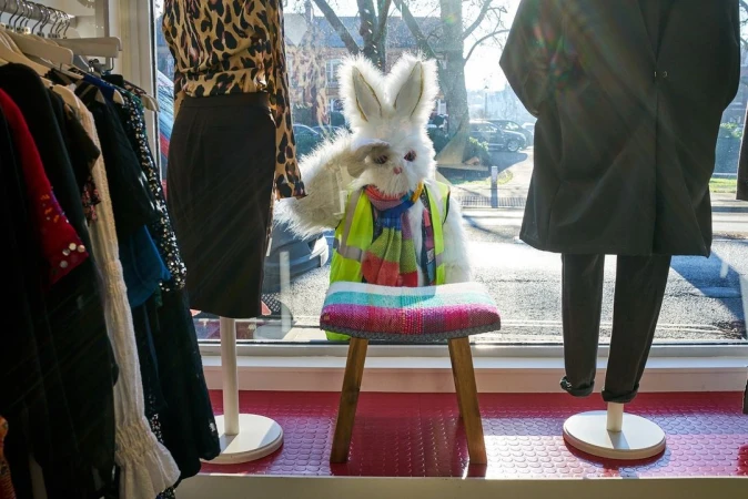 Window display of a large white rabbit figure in safety vest seated on stool with clothing on mannequins and a view to street outside.