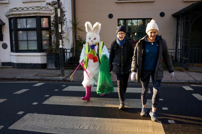 Three people cross a city street on a zebra crossing, including a person in a full white rabbit costume with pink boots and accessories.