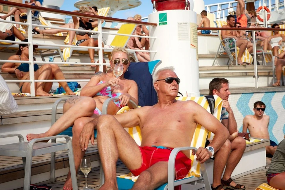 Adults relax in lounge chairs on the deck of a cruise ship under sunny skies, with a clear blue sky in the background.