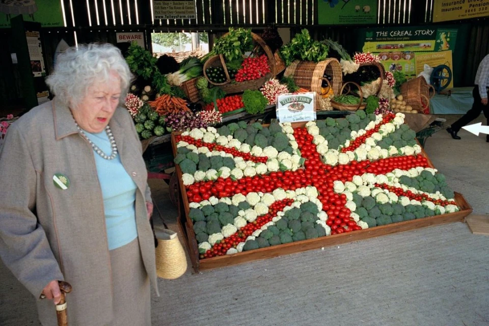 Elderly woman with cane stands near a Union Jack flag made of vegetables at a rural market, illuminated by natural light indoors.