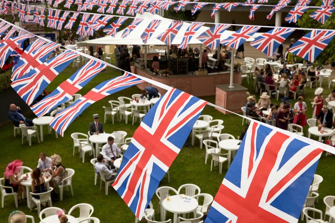 Outdoor social gathering with people sitting at tables on the lawn, adorned with Union Jack flags creating a festive atmosphere.