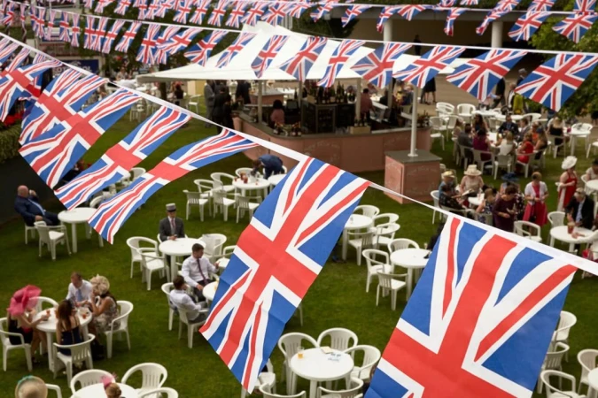 Outdoor social gathering with people sitting at tables on the lawn, adorned with Union Jack flags creating a festive atmosphere.