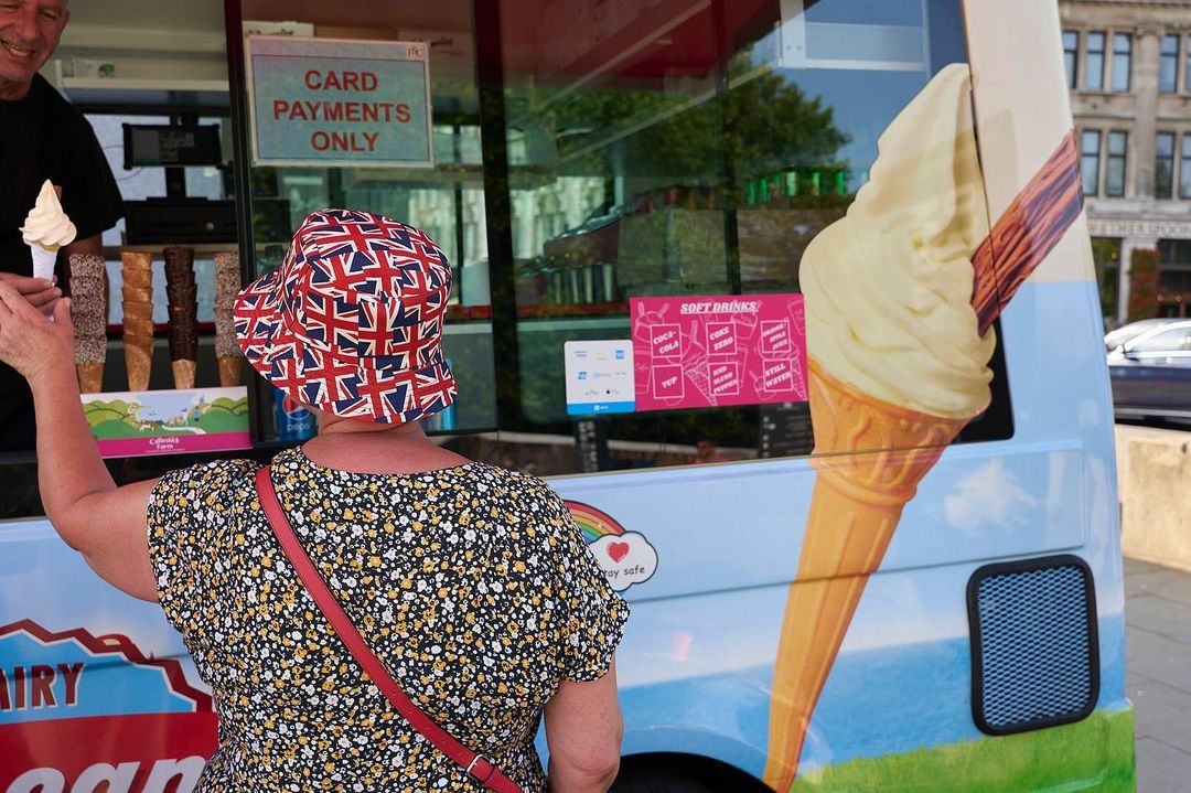 Woman in Union Jack hat buys ice cream from a vendor at an ice cream truck on a sunny day in a city.