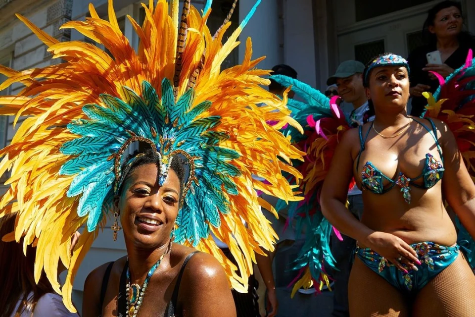 Two women in elaborate feathered carnival costumes smile at the camera during a daytime outdoor event with building in background.