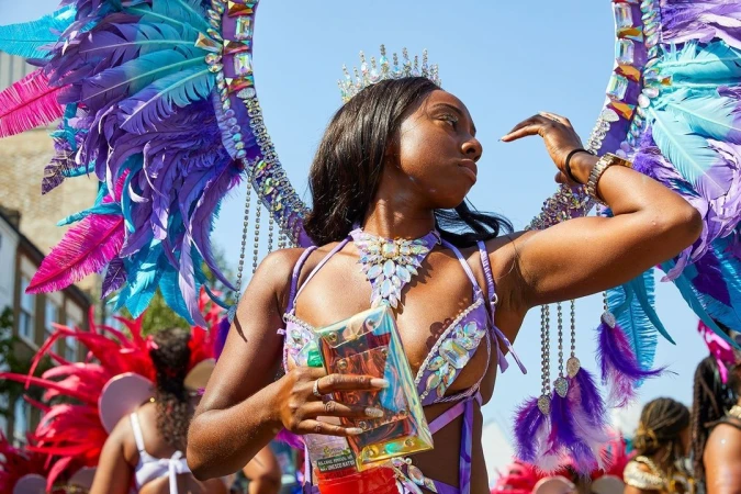 An adult female poses in an outdoor setting, adorned with a vibrant purple and pink feathered costume and jeweled crown and necklace.