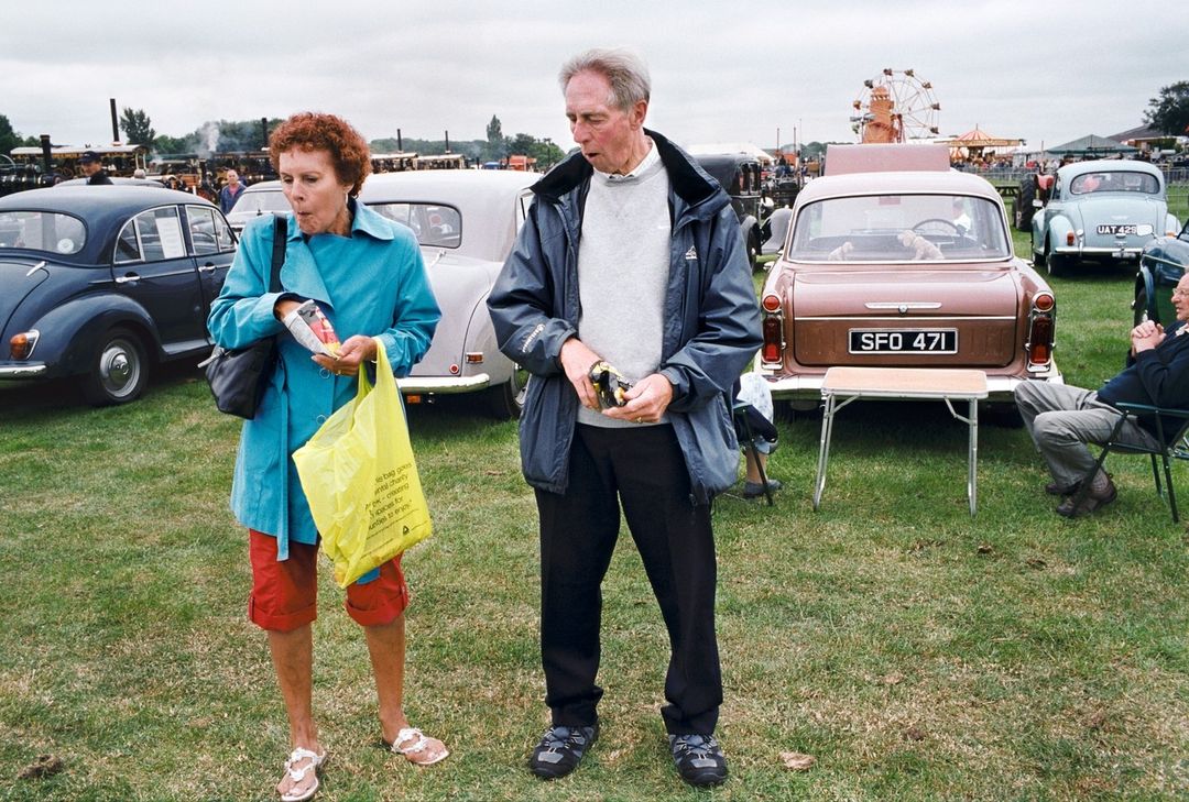 Two adults stand on a grassy field eating snacks in front of vintage cars, with a Ferris wheel in the distance on a cloudy day.