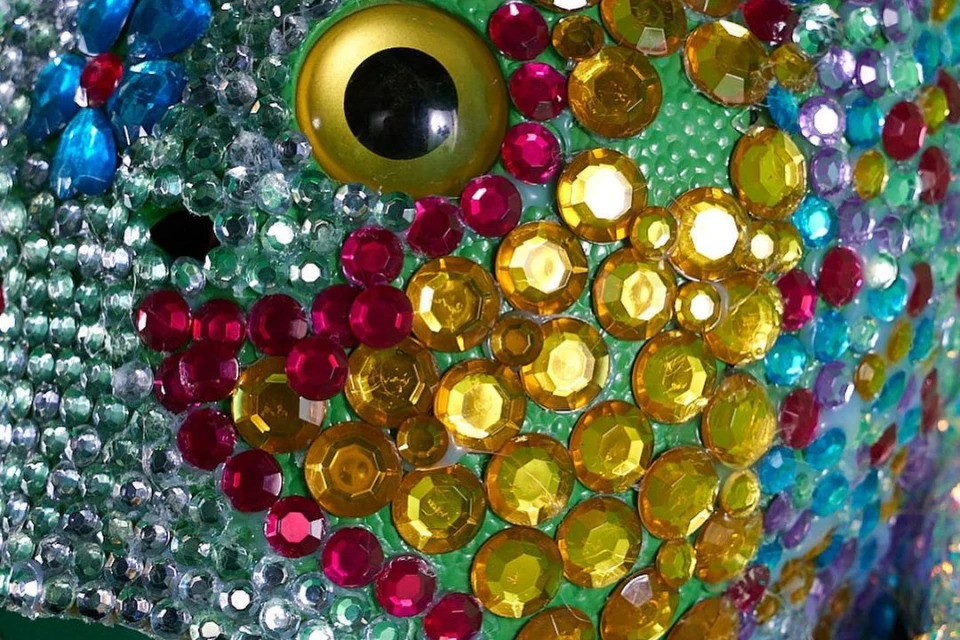Close-up of an abstract, colorful art object resembling a fish covered in bright gems and jewels with a green background and gold eye.