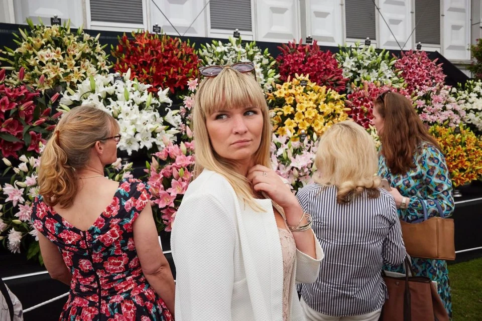 Adult female stands in front of a colorful flower display at an outdoor garden event on a sunny day; other people are in background.