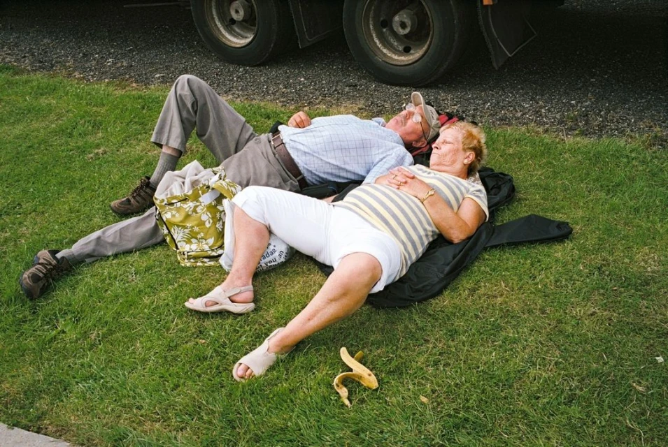 Elderly couple relaxes on lawn by truck. Man and woman are lying down, enjoying the outdoors in daylight with grass and vehicle visible.